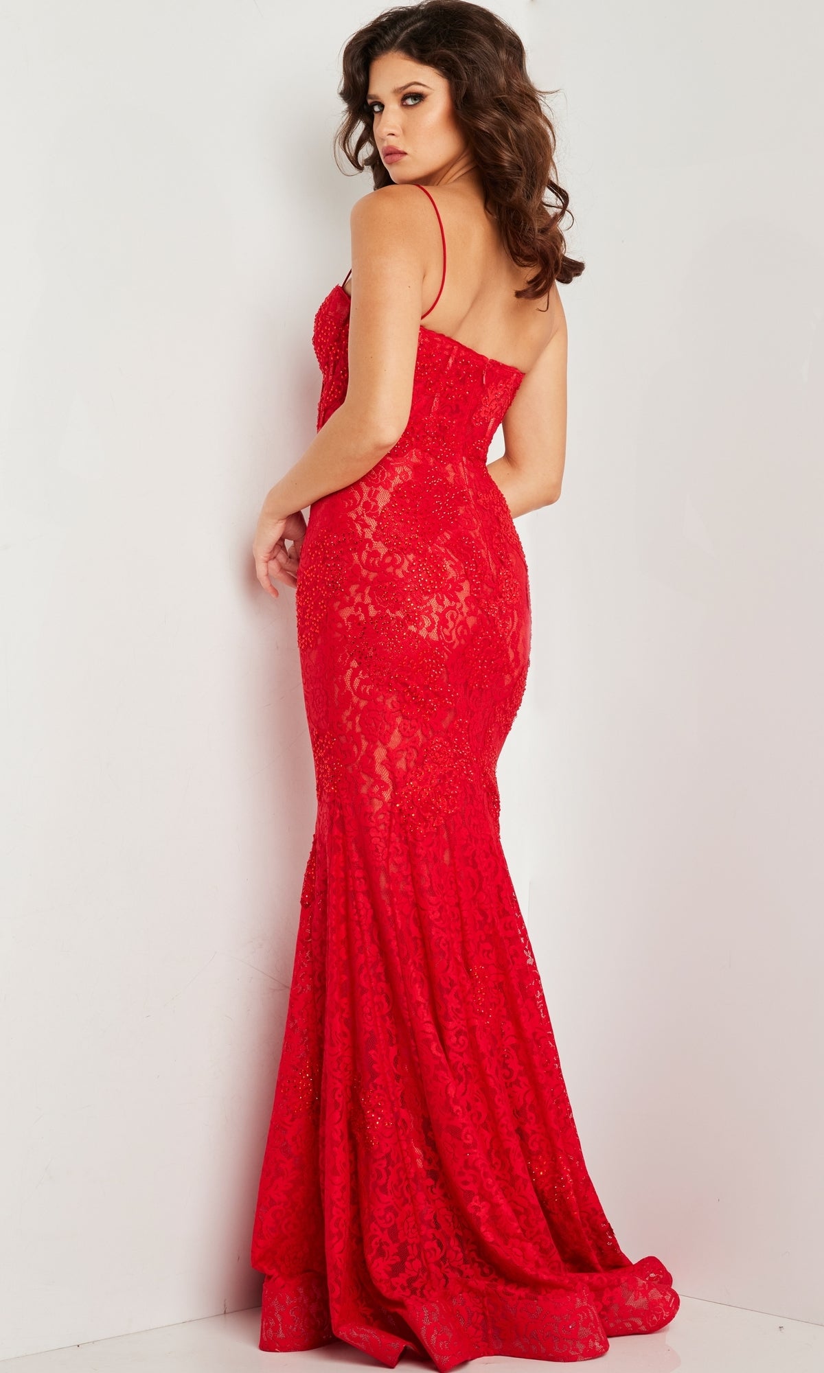 A woman faces away, wearing the fitted Jovani Floor-Length Glitter-Lace Prom Dress 07499 with thin straps, a mermaid skirt, and a slight train. The red prom gown creates a striking silhouette against a plain white background.